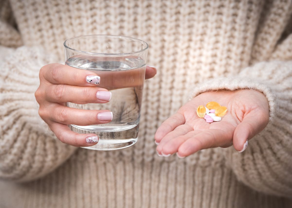 Close-up of a woman's hands holding a glass of water in one hand and a variety of vitamins and supplements in the other, emphasizing the importance of daily health routines. She wears a cozy beige sweater, highlighting a calming and nurturing environment. Miraculous Health offers the finest vitamins and supplements to fuel your health and wellness journey. Visit MiraculousHealth.org to learn more!