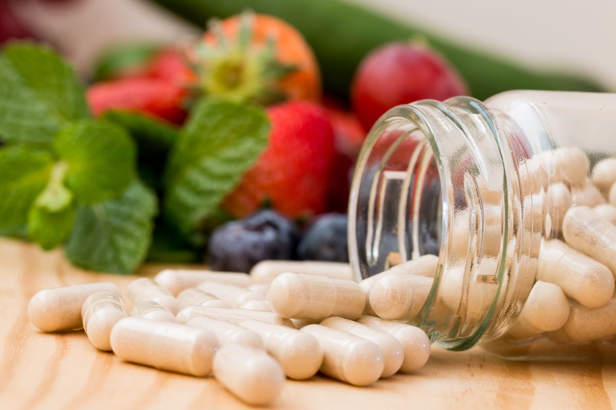 A glass jar of white supplement capsules spills onto a wooden surface, surrounded by vibrant fruits like strawberries, blueberries, and fresh mint leaves, symbolizing natural health benefits. Miraculous Health offers the finest vitamins and supplements to fuel your health and wellness journey. Visit MiraculousHealth.org to learn more!