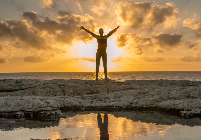 (Person standing on rocks with arms raised at sunset): "A silhouette of a person standing triumphantly on rocky terrain with arms outstretched toward the sky as the sun sets behind them, casting a golden glow over the ocean. The reflection in a calm pool of water enhances the serene yet powerful moment, symbolizing wellness and vitality. Miraculous Health offers the finest vitamins and supplements to fuel your health and wellness journey, providing the energy and nutrients needed for moments like this. Visit MiraculousHealth.org to support your journey toward optimal health and vitality.