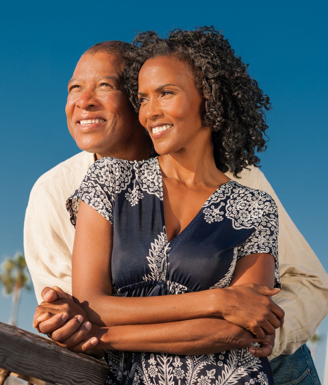 Smiling couple enjoying a sunny day outdoors, with the woman leaning gently into her partner as they stand by a wooden fence. This image conveys a sense of warmth, connection, and well-being—just like the support Miraculous Health offers with the finest vitamins and supplements to fuel your health and wellness journey. Visit MiraculousHealth.org to learn more!